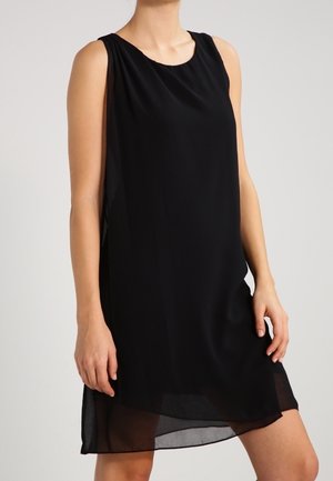 Kobieta ubrana w czarną sukienkę bez rękawów do kolan z przezroczystym dołem, stojąca na tle jasnego, jednolitego tła.