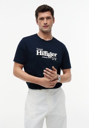 Mies pukeutuneena tummansiniseen Tommy Hilfiger -t-paitaan, jossa valkoinen teksti, ja valkoisiin housuihin, seisoo yksinkertaisen vaalean taustan edessä.
