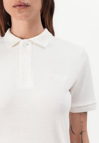 Biely polo tričko vyrobené z textúrovanej bavlny, s trojitém gombíkovým zapínaním a vyšitým logom značky na ľavej strane.