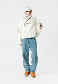 Chaqueta de forro polar blanca con cremallera, capucha acolchada color beige, pantalones de mezclilla azules, parches de acento marrones y botas color beige. Presenta patrones texturizados.