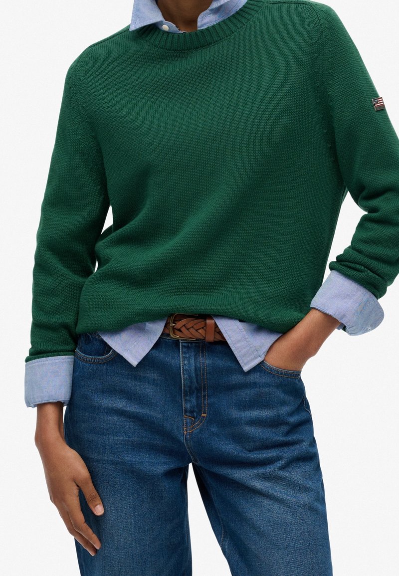 Personne portant un pull en tricot vert par-dessus une chemise bleu clair boutonnée avec les manches retroussées, associé à un jean bleu et une ceinture marron tissée.