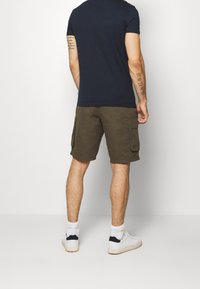 Man som bär en marinblå t-shirt, olivgröna cargo-shorts, vita strumpor och vita sneakers, står vänd mot en vit vägg med synliga arm-tatueringar.