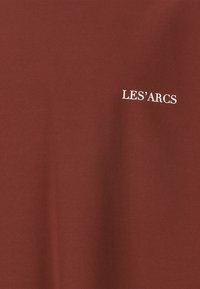 Vinröd T-shirt tillverkad av mjukt tyg med vit text "LES’ARCS" på framsidan, med en enkel design och slät textur.