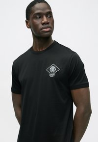 Musta lyhythihainen T-paita sileästä kankaasta, jossa vasemmassa rintataskussa valkoinen, timantinmuotoinen kirjailtu logo.