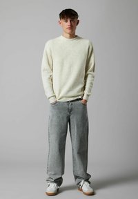 Jaunas vyras stovi su rankomis kišenėse, apsirengęs kreminės spalvos raštuotu megztiniu, laisvais pilkais džinsais ir gerais baltomis sportbačiais pilkoje fone.