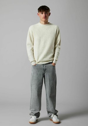 Junger Mann steht mit Händen in den Taschen, trägt einen cremefarbenen gerippten Pullover, lässige graue Jeans und weiße Sneakers vor einem grauen Hintergrund.