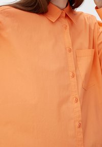 Orange skjorte med knapper lavet af let materiale. Har en spids krave, brystlomme og subtil tekstur med matchende knapper.