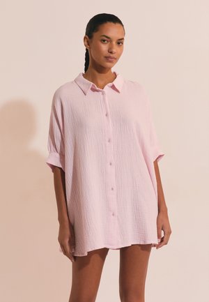 Kobieta z zaplecionymi włosami, ubrana w luźną, jasnoróżową sukienkę koszulową zapinaną na guziki, stojąca na tle bladego tła.