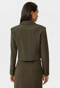 Blazer vert olive cintré avec un col élégant, des épaules structurées et des poignets à un bouton. Tissu lisse avec un design arrière épuré.