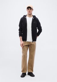 Jeune homme debout portant une casquette beige, un sweat à capuche noir zippé, un t-shirt blanc, un pantalon beige et des baskets noires sur un fond blanc uni.