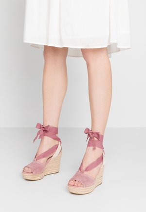 Jambes portant des sandales compensées en suède rose avec des rubans à nouer autour de la cheville, associées à un ourlet de robe blanche juste au-dessus des genoux.