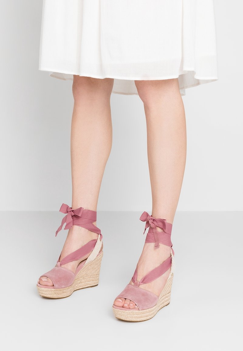 Jambes portant des sandales compensées en suède rose avec des rubans à nouer autour de la cheville, associées à un ourlet de robe blanche juste au-dessus des genoux.