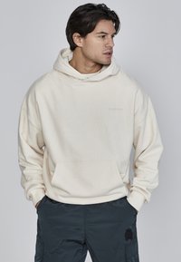 Cremfarbener Hoodie mit einer Fronttasche und gestickten Saumdetails. Kombiniert mit dunklen Hosen, die einen dezenten Logo-Patch haben.