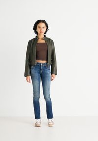 Groene leren bomberjack over een bruine cropped top, gecombineerd met lichtblauwe jeans en beige sneakers. Model staat in een neutrale omgeving.