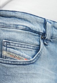 Jeans de mezclilla azul claro con un bolsillo frontal que muestra una etiqueta roja y blanca que dice "Diesel Industry Denim Division." Tejido suave y texturizado.