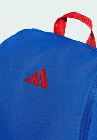 Modrý tkaninový ruksak s červenou vrchnou rukoväťou a červeným logom Adidas so tromi pruhmi na boku. Textúrovaný povrch, obdĺžnikový tvar.
