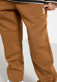 Braune Jogginghose mit gerippten Knieakzenten, goldfarbener Stickerei und einem kleinen Etikett mit der Aufschrift "Play All Day" am Oberschenkel. Hergestellt aus weichem Baumwollmaterial.