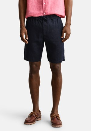 Mann trägt marineblaue Shorts mit Kordelzug, rosa kurzärmeliges Hemd mit Knöpfen und braune Leder-Bootsschuhe, steht vor weißem Hintergrund.