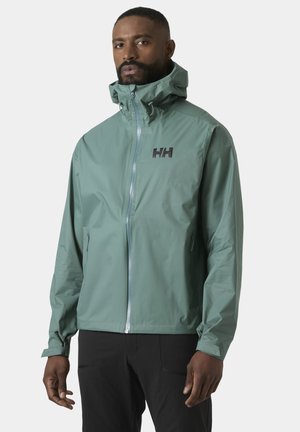 Mężczyzna w zielonej, z kapturem, przeciwdeszczowej kurtce z przednim zamkiem oraz czarnych spodniach, stojący na tle jasnego, jednolitego tła.