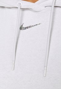 Vit huvtröja i mjukt tyg med en flerfärgad glittrig Nike-logga, med dragsnören och strukturerad yta.