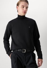 Black turtleneck sweater made of soft fabric, featuring long sleeves and a small logo on the chest, paired with black trousers and a belt.