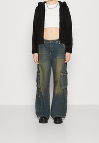 Felpa nera con zip in pelliccia ecologica, top corto bianco, pantaloni cargo in denim blu oversize con tasche, effetto usurato e stivali neri chunky.