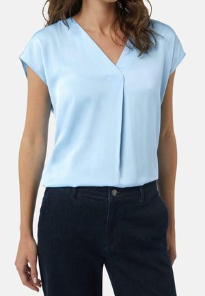 Femme portant un chemisier bleu clair à manches courtes en V, rentré dans un jean bleu foncé avec bouton et passants pour ceinture.