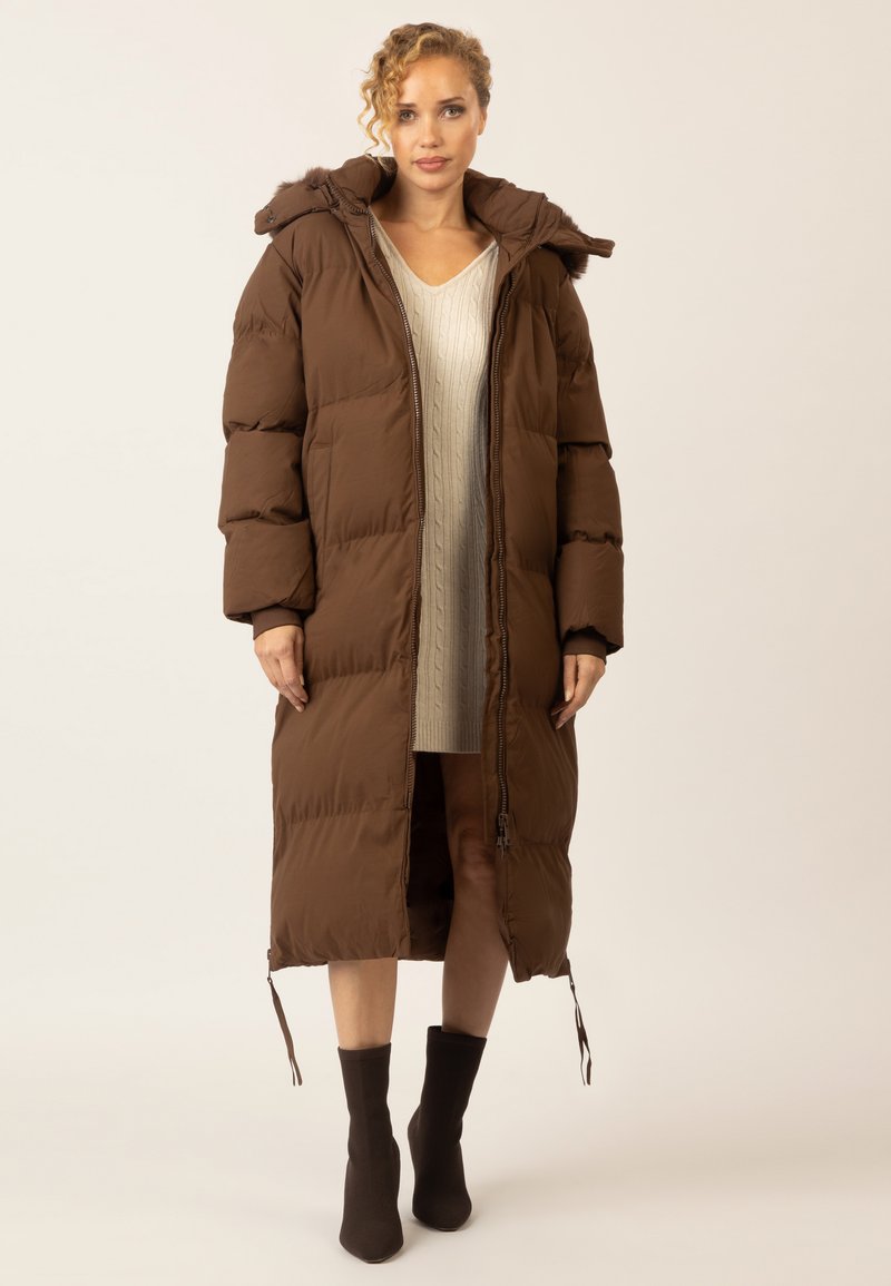 Brauner gepolsterter langer Mantel mit Kapuze, mit einem Reißverschluss, strukturiertem Stoff und Kordelzug am Saum. Wird über einem leichten Strickkleid getragen.