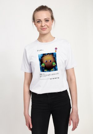 Kvinde iført en hvid t-shirt med en farverig tegneseriefigur og japansk tekst, stående mod en ensfarvet hvid baggrund.
