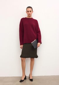 Burgundinės mezginiai megztinis, alyvuogių žalios spalvos fringinė sijonas, juodi bateliai su blokuotu kulnu. Laiko juodą delninę su rožinės aukso akcentais.