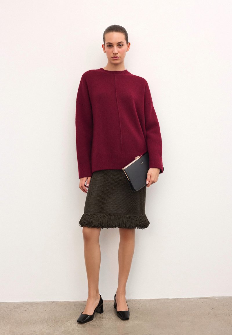 Burgundinės mezginiai megztinis, alyvuogių žalios spalvos fringinė sijonas, juodi bateliai su blokuotu kulnu. Laiko juodą delninę su rožinės aukso akcentais.