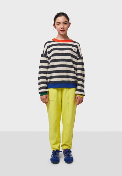 Suéter de punto a rayas en azul marino y crema con cuello naranja; combinado con pantalones de chándal amarillos brillantes que tienen bolsillos y zapatos azules.