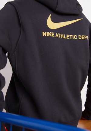 Sort hættetrøje med gul Nike-swoosh og teksten "NIKE ATHLETIC DEPT." på ryggen, båret af en person, der læner sig op ad en blå gelænder.