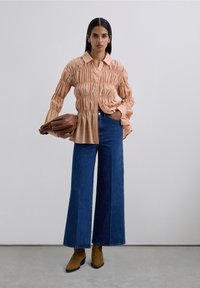 Kvinna iklädd en rynkad beige blus, högmidjade vida jeans i blått, bruna ankelskor, som håller en stor brun clutchväska.