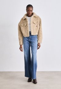 Beige kunstpels cropped jakke med to brystlommer, knappelukking, brukt over en hvit topp og kombinert med vide blå jeans og brune støvler.