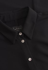 Blusa nera con colletto, caratterizzata da una superficie liscia, con due chiusure a bottone e un'etichetta con la scritta "leschöi" e "Stoccolma."