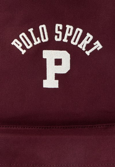 Λευκό κεντημένο κείμενο που λέει "POLO SPORT" πάνω από ένα μεγάλο γράμμα "P" σε βουργουνδί υφασμάτινη υφή.