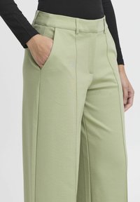 Grüne weite Hose aus glattem Stoff, mit vorderen Taschen und Falten am Bund. Teilweise mit einem schwarzen Oberteil gezeigt.