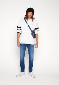 Felpa bianca con cappuccio e dettagli neri, jeans blu e sneakers bianche. Una piccola borsa a tracolla blu con un design a fantasia è indossata attraverso il corpo.