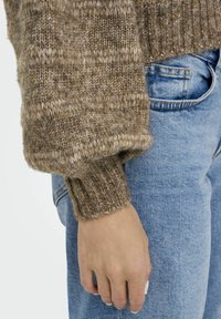 Pull en laine marron avec une finition texturée et douce et des poignets côtelés, associé à un jean en denim bleu clair. Main posée sur la taille.