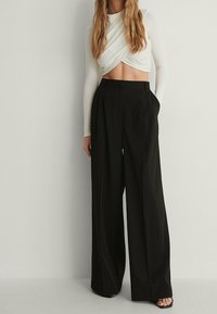 Weißes langärmliges Crop-Top mit verdrehtem Vorderteil, kombiniert mit weiten schwarzen Hosen aus glattem Stoff mit entspannter Passform.