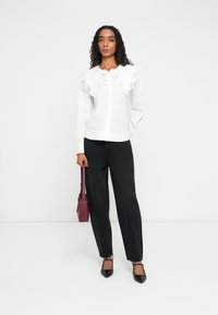 Donna in piedi contro uno sfondo bianco, indossa una blusa bianca con colletto in pizzo, jeans neri, tacchi neri e tiene in mano una borsa bordeaux.