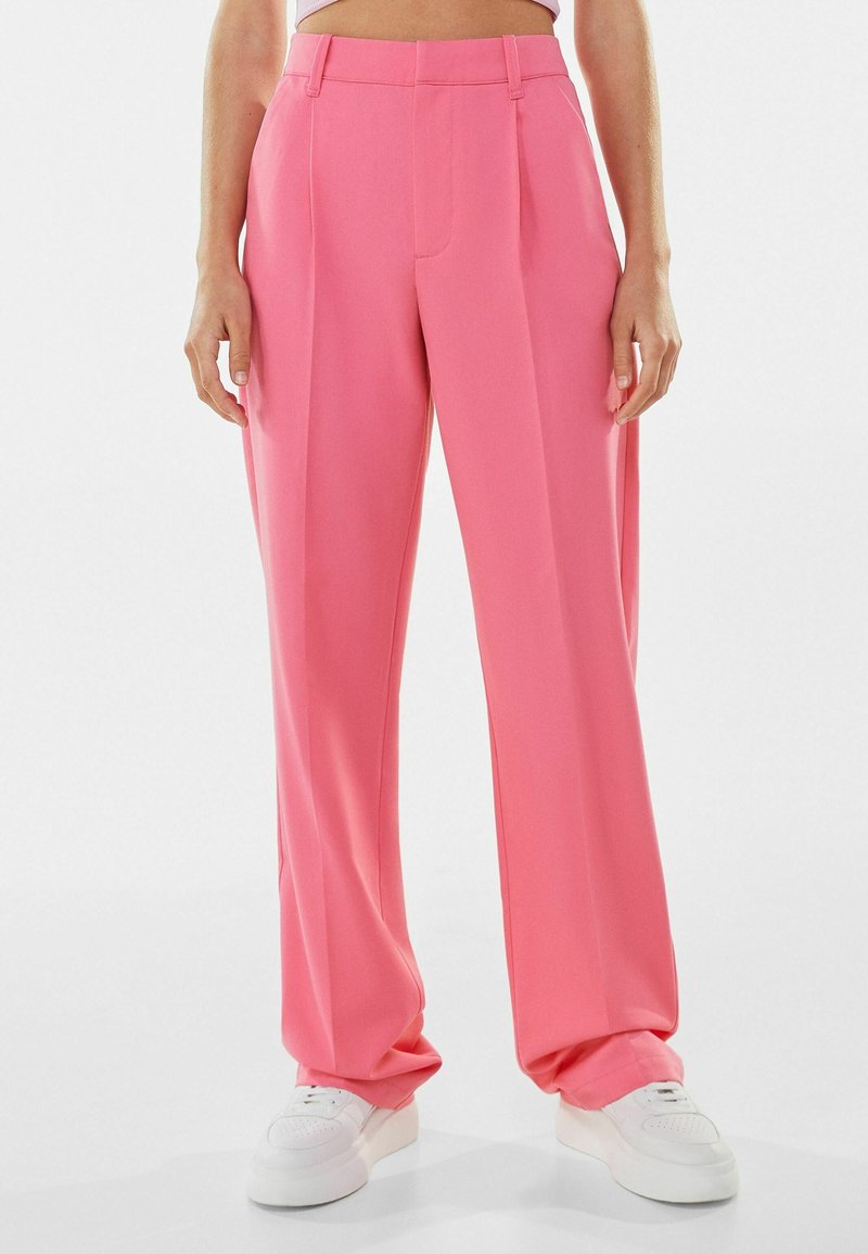Pantalon taille haute à jambes larges en tissu rose vif, avec des plis sur le devant et une texture lisse. Associé à des baskets blanches.