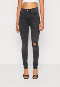 Mujer vestida con jeans ajustados rotos de tiro alto en negro, top cortado blanco, tacones negros de punta abierta, de pie frente a un fondo blanco liso.
