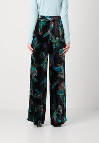 Pantaloni negri cu picioare ample, confecționați dintr-un material moale, având modele colorate cu pene în nuanțe de verde, albastru și violet. Vedere din spate.