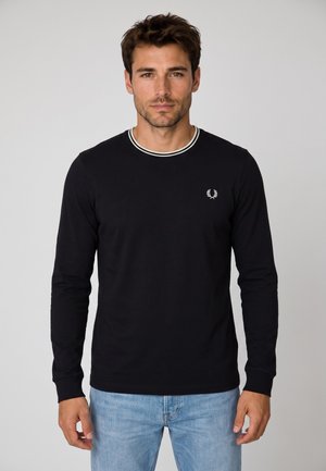 Uomo con capelli scuri che indossa una camicia nera a maniche lunghe con bordino bianco sul colletto e jeans azzurri, in piedi davanti a uno sfondo chiaro uniforme.
