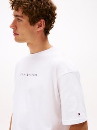 T-shirt bianco in cotone con il logo "TOMMY HILFIGER" sul petto e un piccolo accento del logo sulla manica. Maniche corte, vestibilità rilassata.