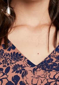 Kvinnas nacke och övre bröst i en rosa och marinblå blommig V-ringad topp och en enkel silverdroppörhänge på vänster sida.