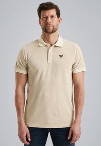 Beżowa koszulka polo z krótkim rękawem, zapięcie na trzy guziki oraz logo motyla na lewej piersi. Wykonana z miękkiej bawełny i dopasowanego kroju.