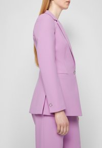 Přizpůsobený lilákový blazer s úzkým střihem, klopovými límci a uzavíráním na jeden knoflík. Disponuje klopovou kapsou a stříbrnými kovovými detaily.